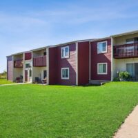 Two-story apartment building with red siding, balconies, and patios, surrounded by a well-maintained green lawn under a clear blue sky.