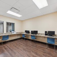A computer lab with three workstations, blue chairs, desktop computers, and a printer on a counter against light-colored walls and a wood floor.