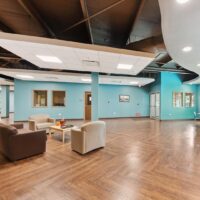 Modern lobby with wood flooring, teal walls, and a seating area with armchairs and a sofa. Large windows and ceiling lights provide ample natural and artificial light.
