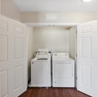 A laundry closet with double doors open, revealing a white top-loading washing machine and a matching dryer on a wood floor.