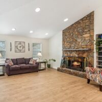 Living room with a stone fireplace, a purple sofa, a patterned armchair, light wood flooring, and two windows with blinds.