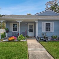 Single-story blue house with white trim, a small covered porch, and a well-maintained lawn with flowers and outdoor seating.