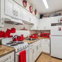 A compact kitchen with white cabinets, brown countertops, and red decor accents including a kettle, oven mitts, microwave, and apple-themed wall art.