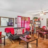 A living and dining area with a black sofa, red and white decor, a wooden dining table, and bright red curtains on a window. The room has hardwood floors and a ceiling fan.