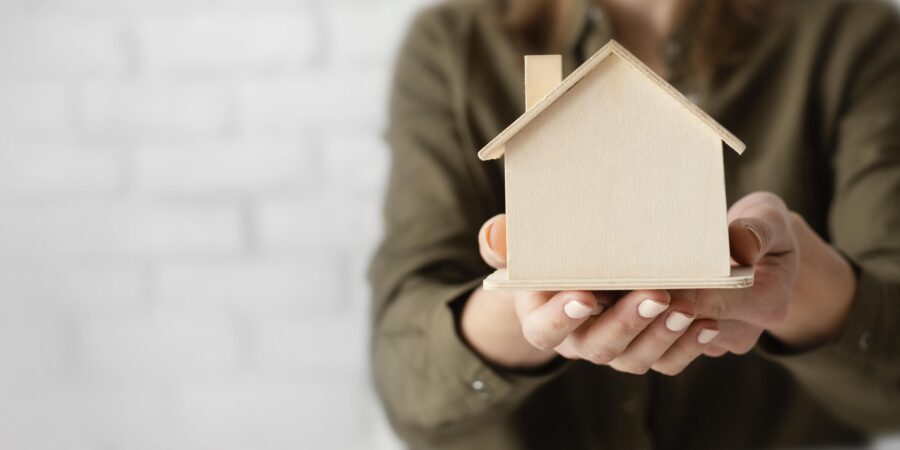 A person with light-colored nail polish holds a small wooden house model in both hands against a blurred white background.