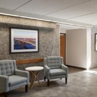 Two gray armchairs and a small round table are arranged against a textured wall, which displays a framed photo of a bridge in a well-lit hallway.