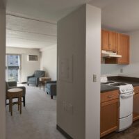 View of a small apartment with a kitchen area featuring a stove and cabinets, and a living area with chairs, small tables, and a window letting in natural light.