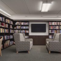 A modern library room with patterned armchairs facing a wall-mounted TV, surrounded by shelves filled with books.