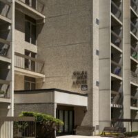 A beige concrete apartment building features balconies and an entrance labeled "Golda Meir House" beside a small bench and shrubbery.