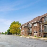 A brick apartment building with balconies is shown beside an empty parking lot under a clear blue sky. Trees line one side of the building.