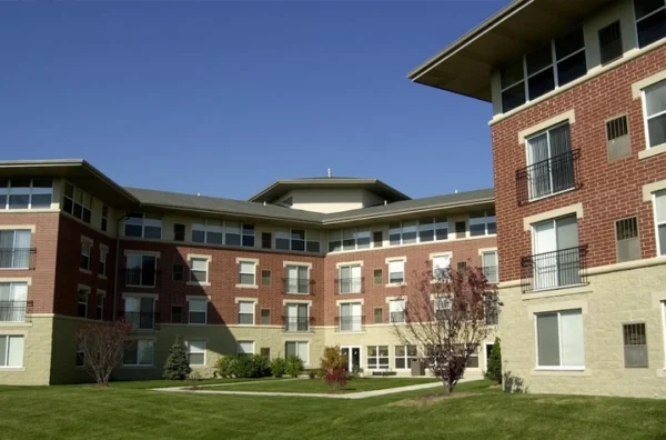 A modern, multi-story apartment building with red brick and beige stone exterior, large windows, and a landscaped green lawn under a clear blue sky.