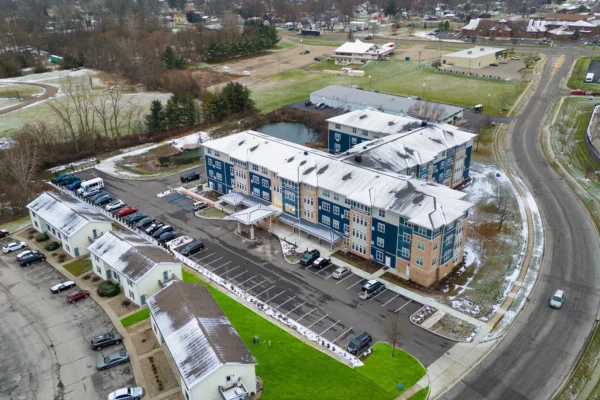 Aerial view of a blue and beige multi-story building with a snow-dusted roof, surrounded by parked cars, roads, grass, and nearby smaller buildings.