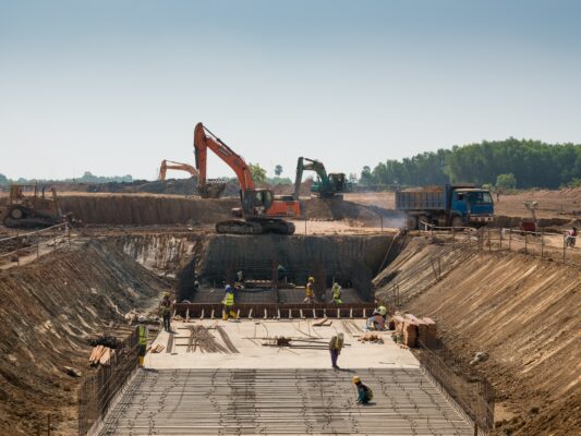 Construction site with workers, excavators, and trucks; workers are laying steel reinforcement while machinery operates in the background.