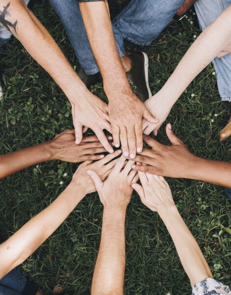 A group of people sitting in a circle outdoors put their hands together in the center over grass, showing unity and teamwork.