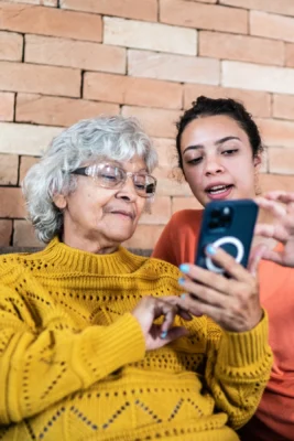 Older woman in a yellow sweater looks at a smartphone while a younger woman beside her points at the screen, both seated in front of a brick wall.