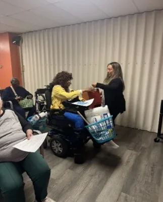 A woman in a wheelchair receives supplies from another woman in a room with other people present.