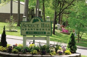 A sign reading "Spruce Hill Apartments" with a phone number stands among flowers and greenery in front of residential buildings and trees.
