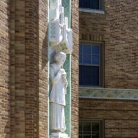 A stone statue of a robed figure holding a scroll is set into a decorative niche on a brick building facade with geometric patterns.