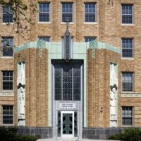 Art Deco-style brick building entrance with geometric metal doors, religious statues on either side, and a sign above the door reading “Saint Charles Senior Living.”.