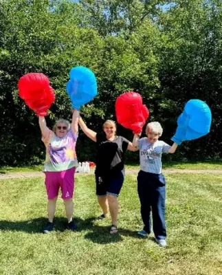 Three people stand on grass holding up large inflatable red and blue boxing gloves, with green trees in the background on a sunny day.