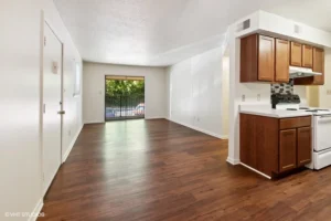 Unfurnished apartment interior with wood flooring, white walls, a kitchen with brown cabinets, and a sliding glass door leading to a fenced outdoor area.