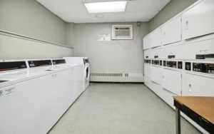 A small laundry room with multiple white washing machines and dryers lined up against two walls, an air conditioning unit, and a wooden table in the corner.