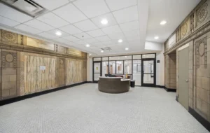 A lobby with patterned walls, tiled floor, a circular reception desk, glass doors, and fluorescent ceiling lights.