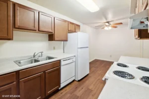 A kitchen with brown cabinets, white countertops, a double sink, dishwasher, refrigerator, electric stove, and wood floor under a ceiling fan light.