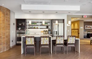 Modern kitchen area with four patterned chairs at a counter, pendant lights above, stainless steel appliances, and open shelving with decorative items.
