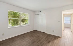 Empty room with light-colored walls, wood-style flooring, a window with blinds, a closed closet with double doors, and a hallway visible to the right.