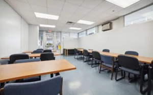 A classroom with several wooden tables and blue chairs arranged in rows, white walls, a tiled ceiling, and large windows letting in natural light.
