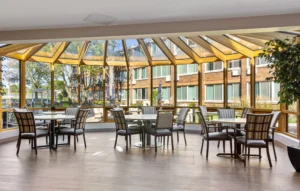 A sunroom with large windows, several tables, and chairs arranged for dining or socializing, overlooking an outdoor courtyard.