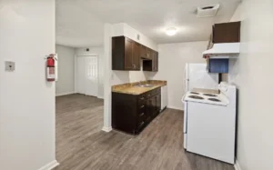 Small kitchen with dark cabinets, white appliances, wood-look flooring, and a fire extinguisher mounted on the wall near an open living area.