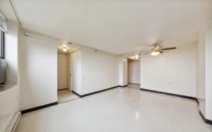 Empty room with white walls, tile flooring, ceiling fan with light, and exposed pipes near the ceiling; doorways lead to other parts of the space.