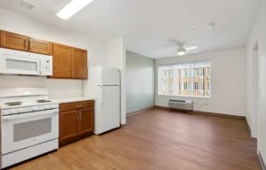 A small apartment kitchen with wooden cabinets, white appliances, and an adjacent open living area with large windows and wood-style flooring.