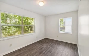 Empty room with white walls, dark wood flooring, a ceiling light, and two windows letting in natural light with a view of greenery outside.