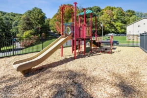 A playground with a tan slide, red climbing structures, and a green-roofed platform, set on wood chips and surrounded by a black fence. Trees and buildings are visible in the background.