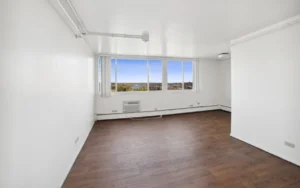 Empty, unfurnished room with white walls, wood-look flooring, large windows showing an outdoor view, and a wall-mounted air conditioning unit below the windows.