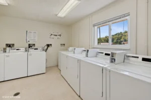Laundry room with multiple coin-operated washing machines and dryers, a window letting in daylight, and instructions posted on the wall.