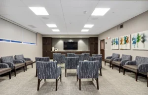 A modern waiting room with patterned chairs arranged in rows facing a large wall-mounted TV, glass tables, abstract artwork, and neutral-toned walls and carpet.