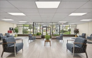 Spacious waiting area with patterned armchairs arranged in a semi-circle, large windows, indoor plants, and bright overhead lighting.