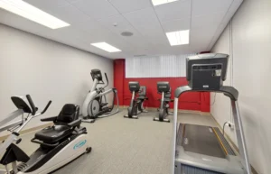Small gym room with a treadmill, two stationary bikes, an elliptical machine, and a recumbent bike, all on a carpeted floor with white walls and a red accent wall.
