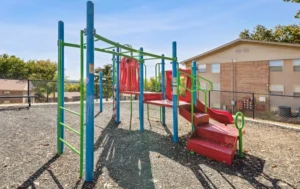 A small outdoor playground with red slides, green and blue climbing bars, on a gravel surface, near a fenced area and brick apartment buildings in the background.