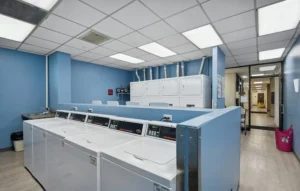 A clean laundry room with top-loading washing machines in the foreground, stacked dryers in the background, blue walls, and overhead fluorescent lighting.