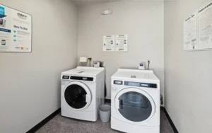 A laundry room with a front-loading washing machine and dryer side by side, instructional signs on the walls, and a small trash can between them.