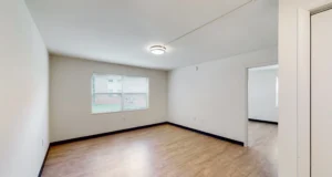 Unfurnished room with light-colored walls, wood flooring, a ceiling light, a window with blinds, and an open doorway to an adjacent room.
