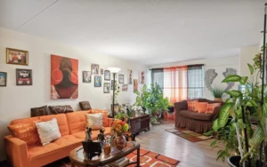 A living room with orange and brown decor, wall art, family photos, potted plants, a sofa, a daybed, and a glass coffee table on a patterned rug.