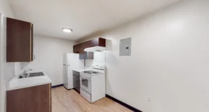 A small kitchen with white appliances, wood cabinets, a refrigerator, stove, range hood, sink, and wood flooring. The walls are white and there is a ceiling light.