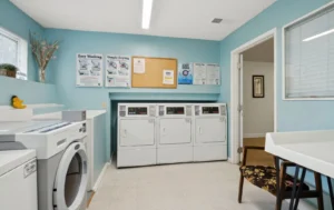 A small laundry room with four front-loading dryers, a washing machine, a bulletin board, blue walls, and informational posters.