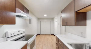 Modern kitchen with brown cabinets, white countertops, an electric stove, and wood flooring, leading to a hallway with double white closet doors.